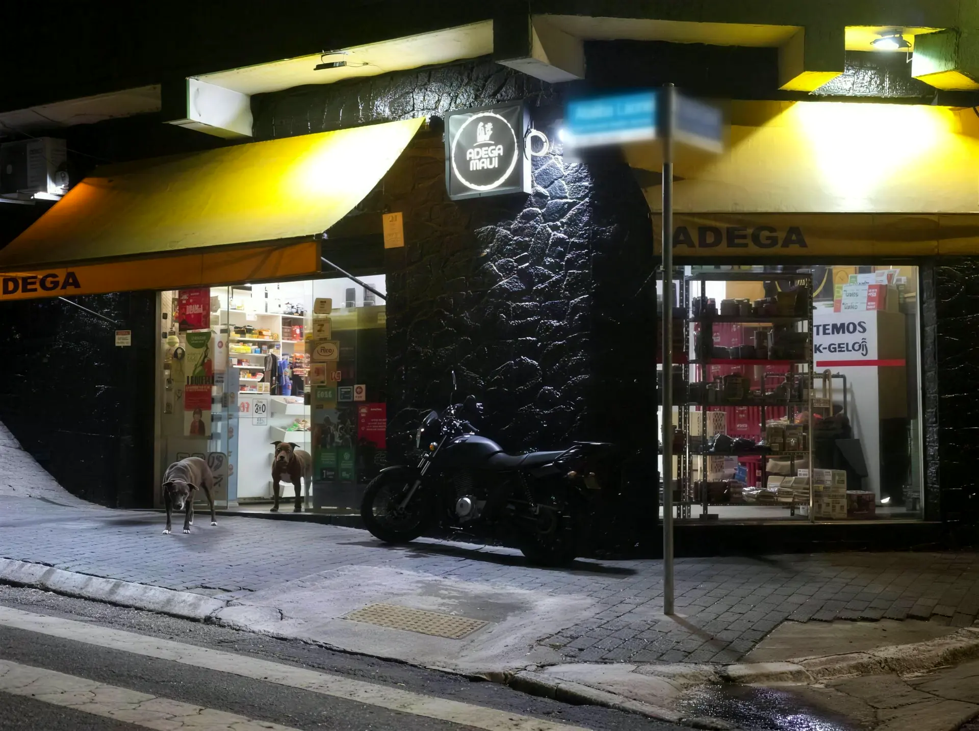 Fachada da Adega Maui à noite na esquina da Av. General Ataliba Leonel: pedra preta, toldo amarelo aceso, placa com o logo, uma moto parada e os cachorros na porta